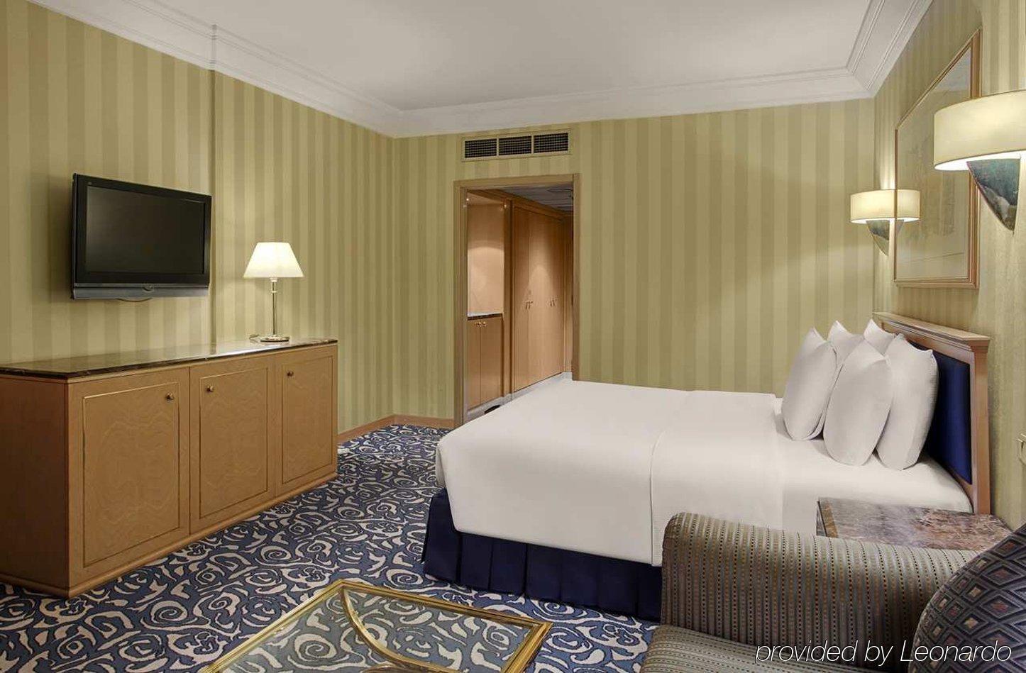 Makkah Hotel La Meca Exterior foto The photo shows a hotel room featuring a double bed with white bedding and several decorative pillows. There is a television mounted on the wall opposite the bed, along with a bedside lamp on a small table. The walls have a striped pattern in light c