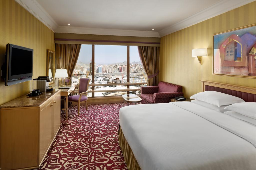 Makkah Hotel La Meca Exterior foto The photo shows a hotel room with a modern and comfortable design. In the center, there is a large bed with white linens, flanked by bedside tables. A plush sofa and a small coffee table are positioned near a large window that provides a view of a ci